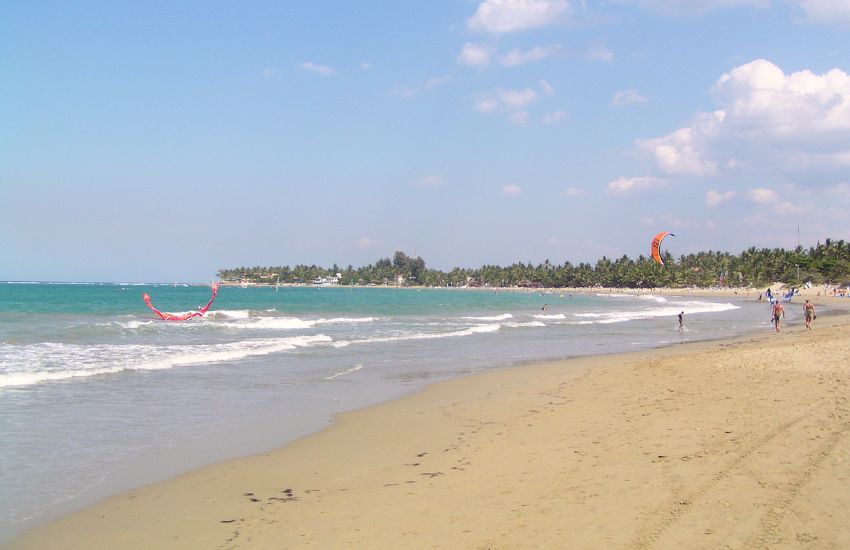 Peščena plaža Cabarete v Dominikanski republiki s palmami, nežnimi valovi in barvitimi kajti v zraku – priljubljen spot za windsurfing in kite surfing.
