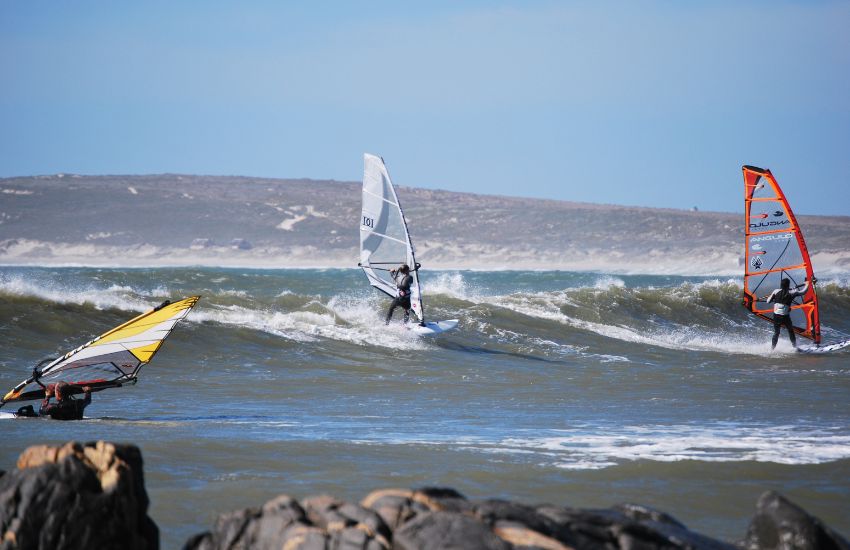 Trije windsurferji na valovih ob obali Cape Towna, s hribovitim ozadjem pod jasnim modrim nebom.