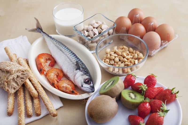 Različna živila, ki pogosto povzročajo alergije, razporejena na mizi &ndash; riba, kozice, jajca, mleko, ore&scaron;čki, ara&scaron;idi, kruh (gluten oziroma celiakija) ter sveže sadje (jagode in kivi).