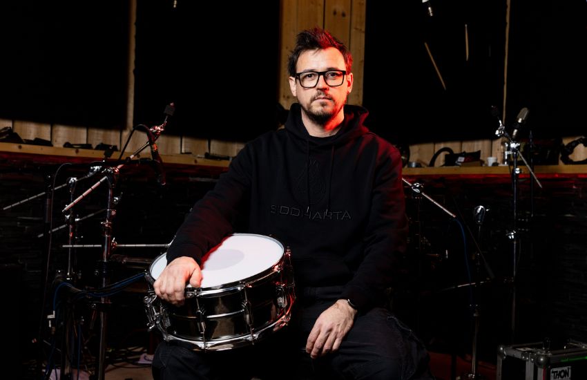 Bo&scaron;tjan Meglič, bobnar skupine Siddharta, sedi v glasbenem studiu in v naročju drži kovinski snare boben in je obkrožen s studijsko opremo.