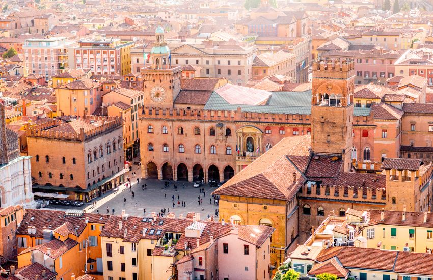 Zračni pogled na trg poln ljudi Piazza Maggiore v Bologni z zgodovinskimi zgradbami iz rdeče opeke, vključno s palačo Palazzo d'Accursio in stolpom z uro.