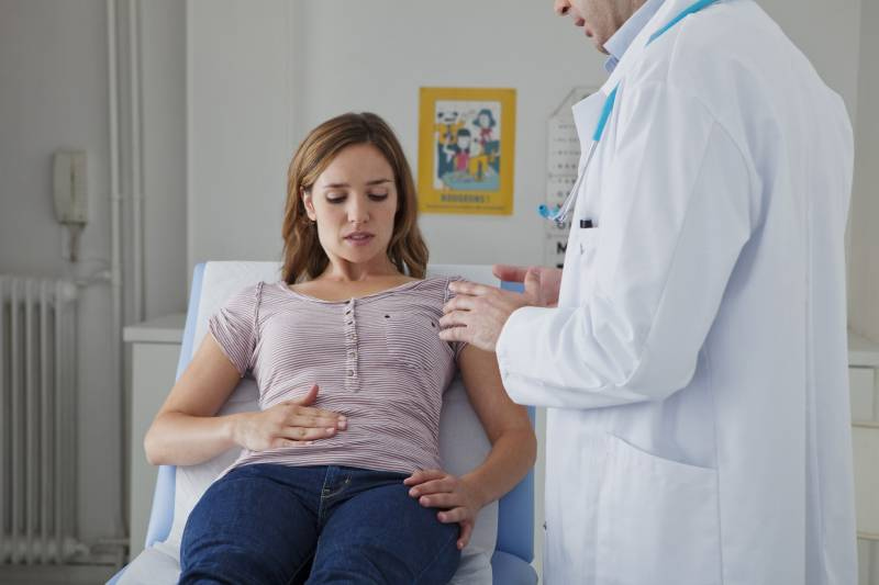 Ženska leži na preiskovalni mizi v ambulanti in se z roko drži za spodnji del trebuha, medtem ko zdravstveni delavec v beli halji stoji ob strani in razlaga postopek preiskave. V ozadju so vidni medicinski plakati in oprema.