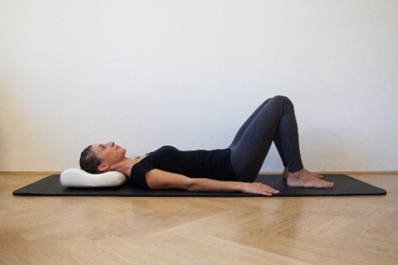 Ženska na hrbtu na telovadni blazini, glavo ima podprto z vzglavnikom, kolena so pokrčena, stopala so postavljena na tla, roke pa spro&scaron;čeno ob telesu.