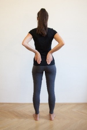 Ženska pokončno stoji na telovadni blazini, s hrbtom je obrnjena proti kameri, noge so v &scaron;irini bokov, dlani pa položene na spodnji del hrbta.