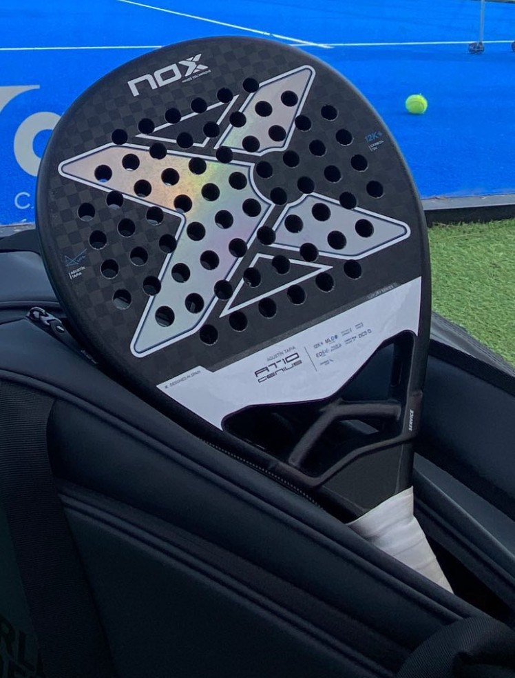 Lopar za padel znamke NOX, naslonjen v &scaron;portni torbi ob modrem padel igri&scaron;ču, z zeleno žogico v ozadju.