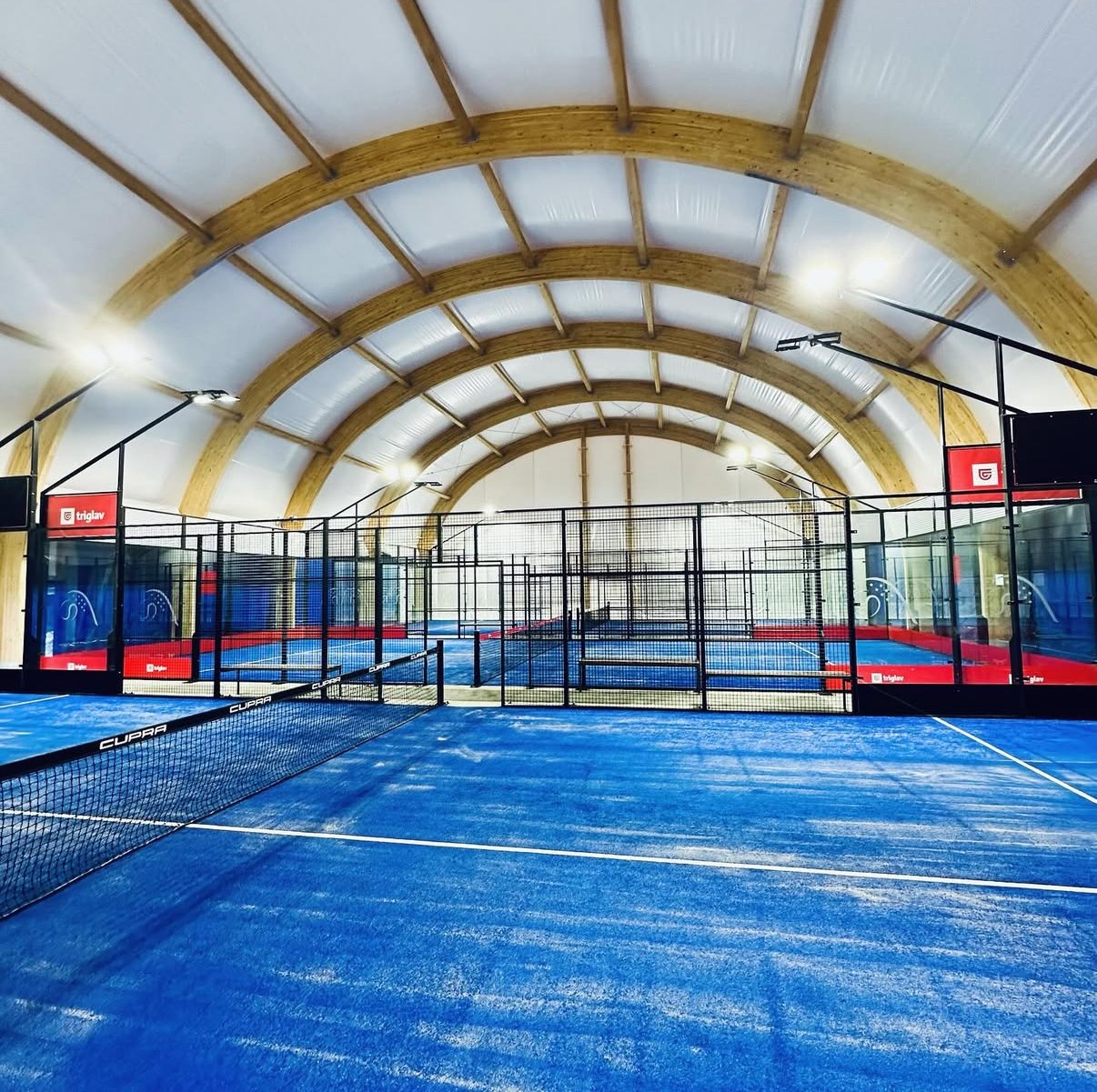 Notranje padel igri&scaron;če z modro igralno povr&scaron;ino, mrežo in steklenimi stenami pod obokano streho &scaron;portne dvorane.