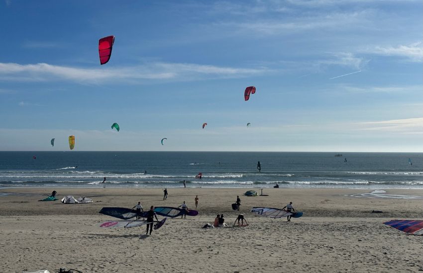 Peščena plaža polna winsurferjev, ki nosijo jadra in deske v morje in pripravljajo windsurfing opremo, v mirnem morju že kite surferji.