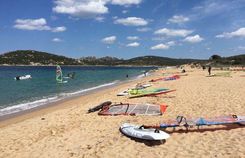 Peščena plaža, na njej razporejena surfing oprema - deska in jadra, v morju windsurferji