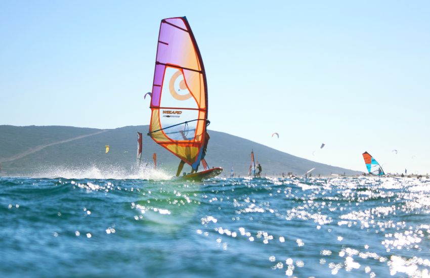 Windsurfer z barvitim jadrom drsi po morski gladini, v ozadju drugi surferji in obala Rodosa v Grčiji