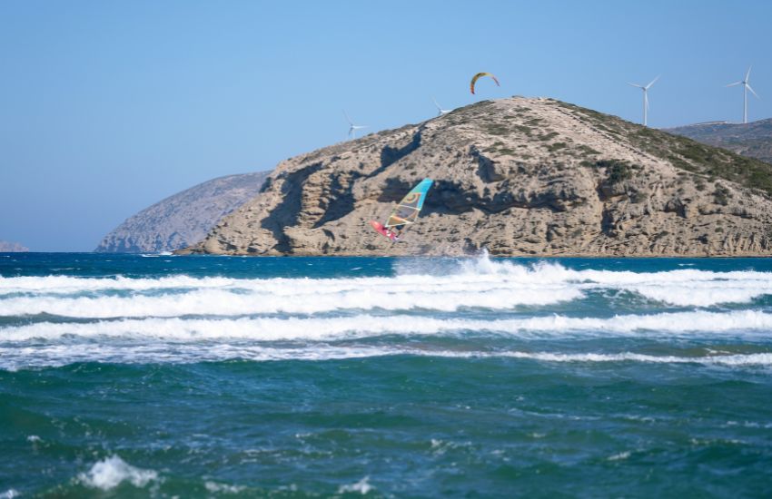 Windsurfer izvaja trik - skok v zraku med valovi, v ozadju tudi kajt in skalnata obala Rodosa