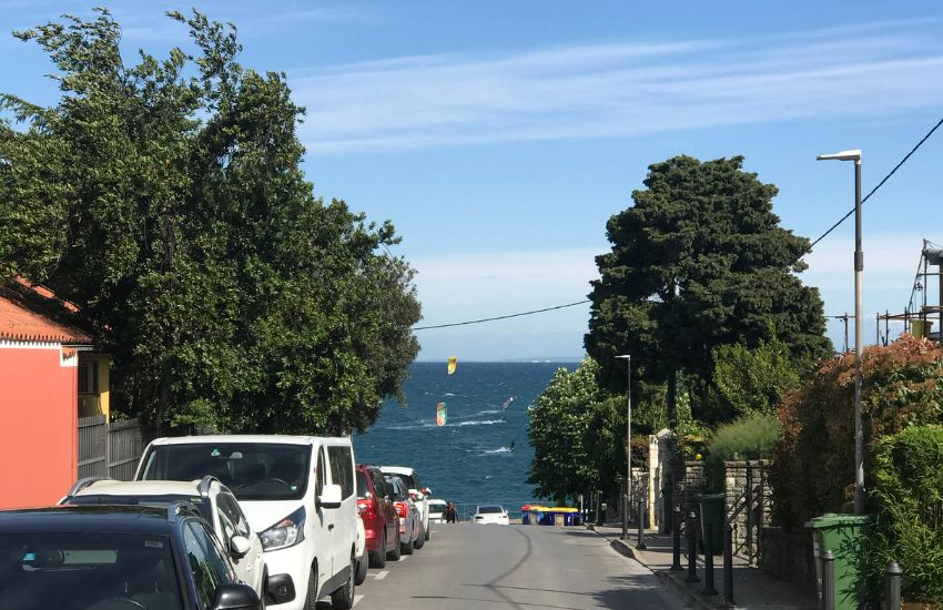 Pogled z vrha ulice, ceste proti morju. Na levi strani ulice so parkirani avtomobili, v daljavi na morju pa so vidna jadra windsurferjev.