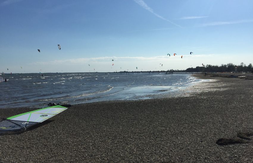 Pogled s plaže na morje z majhnimi spenjenimi valovi. Na plaži leži jadro za surfanje na deski, v ozadju pa vidno veliko kajtov.