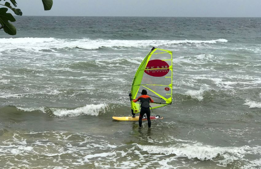 Windsurfer oblečen v neopren z desko z jadrom ob vstopu v vodo na kamniti plaži v Dajli