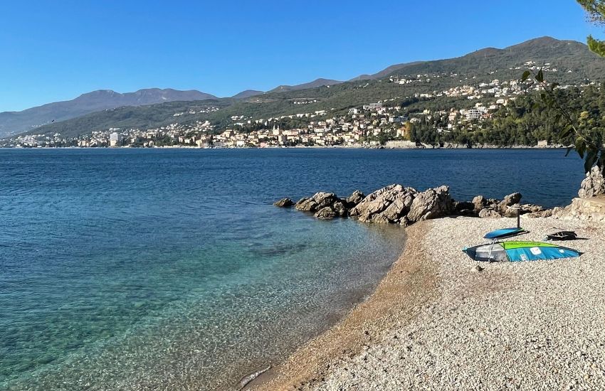Prazna plaža s kamenčki, na kateri leži deska in jadro za windsurfanje, v ozadju obmorsko mesto in zaliv z mirno, čisto vodo.