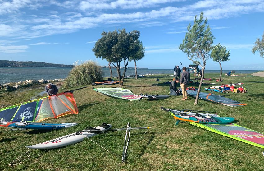 Windsurfi in jadra razprostrta po zelenici na plaži v Seči, v ozadju viden vstop v morje.
