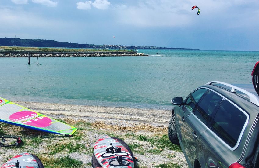 Siv karavan parkiran na plaži tik ob vodi, ob njem položene surferske deske in jadro, v daljavi kite v zraku na morju.
